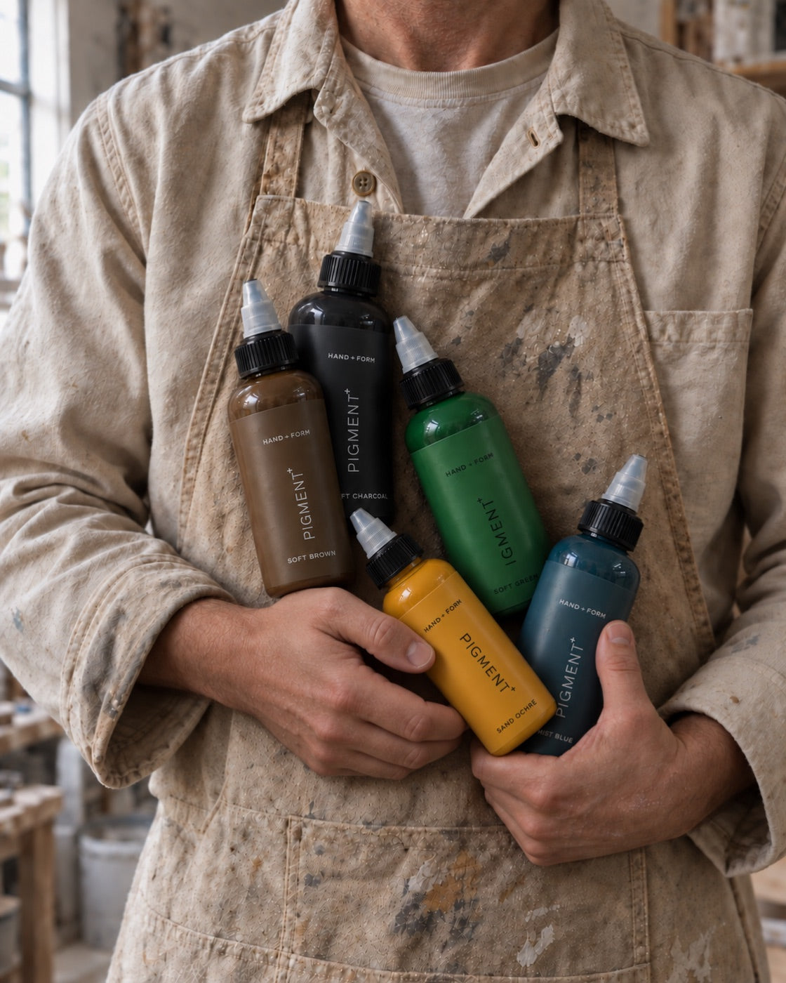 A maker in a studio apron holding the five HAND + FORM Pigment+ bottles